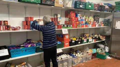 A volunteer is stacking shelves in the food hub store room which shows lots of other food items on the shelves and in crates.