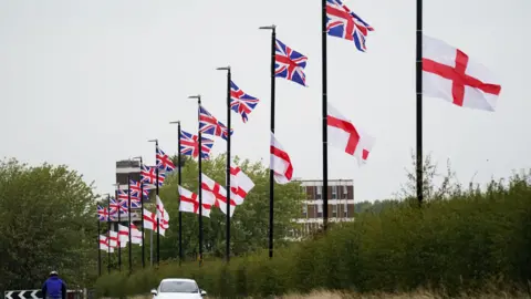A long row of lampposts all have one St Georges flag and one Union flag attached to them. 