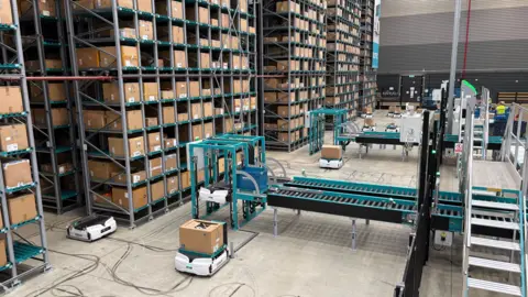 Ben Schofield/BBC Inside the Decathlon warehouse. On left of the image and stretching into the background are rows of shelves with many cardboard boxes each neatly sorted and stored. On the floor in front of the shelves several low-rise white robots can be seen, some are carrying boxes while others are not. There are also industrial-looking staircases and conveyor belts.