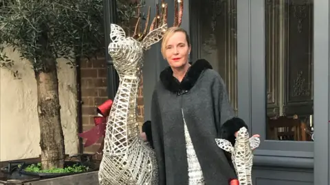 Kirsty Toner A woman wearing a dark grey cape over a white dress poses for a photograph while standing between two metal frame statues of deer.