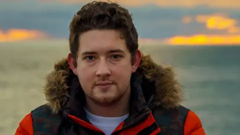 Family A man with thick dark hair and stubble is looking at the camera. He is wearing a red and black jacket with a furry hood