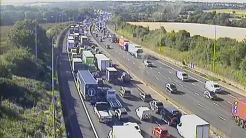A traffic camera's view of a motorway. There is heavy congestion on the left-hand carriageway - full of lorries and cars. The other carriageway is fairly busy, but traffic is moving.
