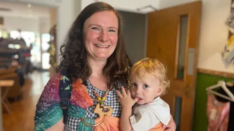 A woman wearing a checked dress and a colourful baby sling is holding her toddler and smiling to the camera. She has long dark brown hair and is standing in a meeting place for parents with young children. 