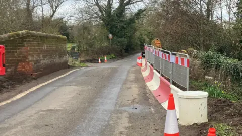 Cones, bollards and fencing along the right hand side of the bridge. Some brickwork damage can be seen. A road going through centre of image. On the left is the undamaged side of the bridge.