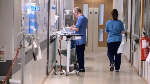 A general view of a hospital corridor at the RAH. A male nurse stands at a computer on wheels, inputting information outside the window into the ward. Another nurse, in a similar blue uniform is seen walking away from the camera towards a double door, carrying a white plastic apron. There are windows on either side of the corridor and boxes of gloves, equipment hanging on the walls.