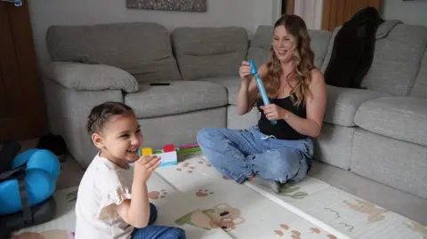 Shaun Whitmore/BBC Amelia and her mum Karis sitting on the floor playing and blowing bubbles. Amelia is sitting down on the left of the picture and she is wearing blue jeans and a light frilly top. Her mum Karis is sitting on the right of the picture with her back against a grey sofa. She is wearing jeans with a black tank top and has a bubble wand in her hand. The pair are blowing bubbles.