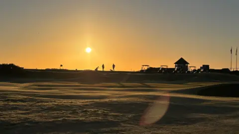 Golfers silhouetted on the course against an orange sunrise glow.