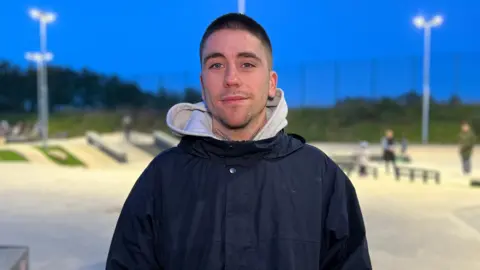 Eddie looks at the camera as he wears a big dark blue coat with a white hoodie on the inside. He is standing in front of a skate part with people practicing behind him. Eddie has short, shaved hair.