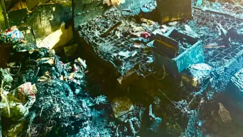 Paul Graham A fire damaged bedroom, with charred remains. There are lots of items around the room, but you cannot make out what they were. There are beds with some springs on them. 