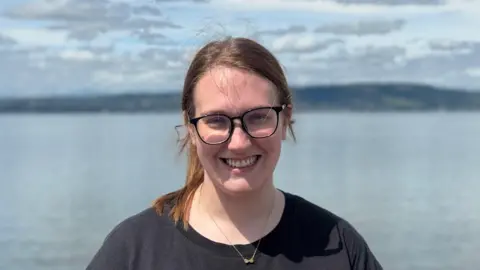 Close up image of Shannon Grimley wearing black t-shirt smiling at camera. She has black framed glasses on and long brown hair tied back in a ponytail. She is standing in front of the open sea with green hills in the far background.