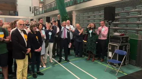 Political candidates with varying colours of rosettes on their clothing standing in a line in a sports hall