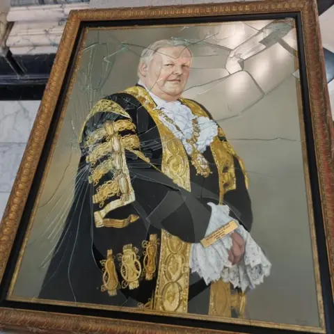Belfast City Council A full-length photo of the damage to a gold-framed oil painting of former Belfast lord mayor Lord Browne.   The painting shows a grey-haired man in a black and gold ceremonial robe, wearing a gold mayoral chain around his neck.   Most of the glass protecting the painting has been shattered. 