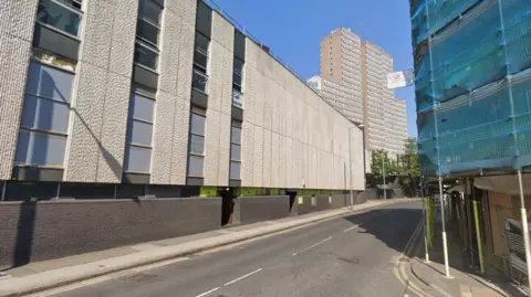 Streetview image of Glasshouse Street in Nottingham, an urban road with a concrete shopping centre on one side and buildings with scaffolding on the other