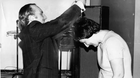 Kenneth Black Mary being presented with a medal by a man wearing a suit. She has short dark hair and is wearing a white top.