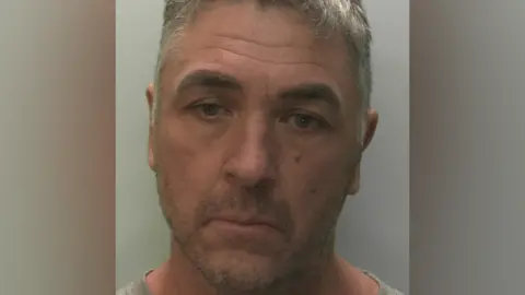 Police mugshot of a man with stubble and greying hair looking down slightly