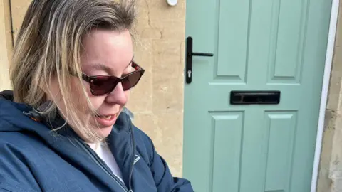 Claire Cavanagh A blonde woman in sunglasses and a dark blue anorak and white T-shirt sits outside a house on the street where the Residents' Parking Zone is due to be brought in. 