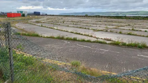 A run down piece of industrial land with tarmac and concrete and grass growing up between the cracks