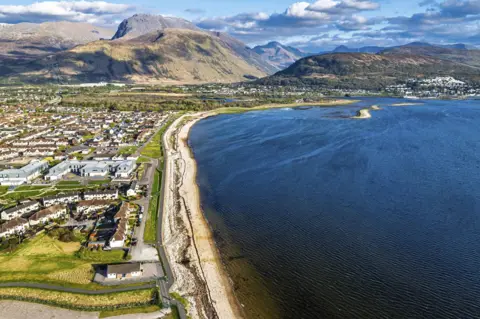 Getty Images An aerial shot showing dozens of houses on a green grassy stretch next to a body of water on the right hand side. In the distance, tall grey and green hills can be seen in front of a cloudy blue sky. 