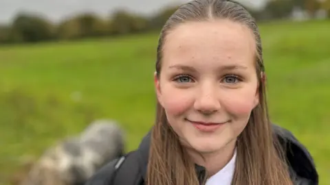 Leah olha para a câmera sorrindo - ela tem longos cabelos castanhos e está vestindo seu uniforme escolar