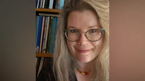 Lisa Brundle A bespectacled woman with long blonde hair posing for a picture. She is wearing a multi-coloured necklace and is standing in front of a bookcase.