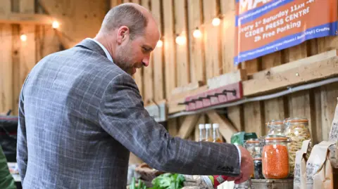 PA Media The Prince of Wales during a visit to the Apricot Centre in Dartington, Totnes. He wears a grey blazer and is looking at products at an indoor market stall, with jars of dry goods, bottles, fresh vegetables and a sign advertising a regular market event.