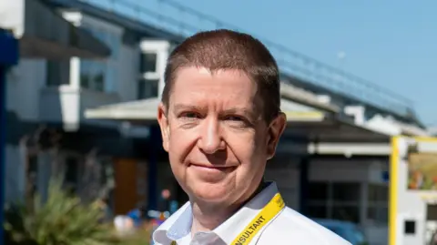 West Suffolk NHS Foundation Trust Dr James Mapstone smiles. He has short thin hair and wears a white shirt with a yellow lanyard around his neck.