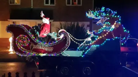 A red sleigh covered in fairy lights withe the Fire Fighters Charity logo on the side a person dressed as Santa waves as it is driven past 