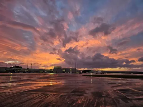 Sam Campbell Vivid sky of orange, yellow, blue and purple colours reflecting on the runway of Glasgow airport.