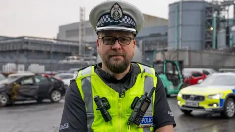 A police officer wearing full uniform which includes a white hat with a black and white checked band. He is wearing a black top with police Scotland written on both sleeves and a high-vis vest with a walkie-talkie and a torch attached to the front. He is standing in a scrap year with a police car in the frame.