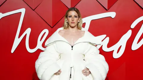 Ellie Goulding wearing an off the shoulder white winter coat. She has dark blonde hair that goes down her back and silver earrings. She is stood in front of a red background with white wording across it. 