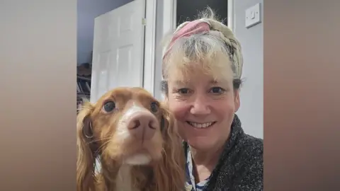 Katie Banfield A picture of Katie Banfield and her dog, Alfie. Katie is smiling and has blonde hair and Alfie is a brown spaniel. The picture is a selfie and Alfie looks slightly off to the side while Katie looks directly at the camera. She is wearing a black, patterned jumper. 