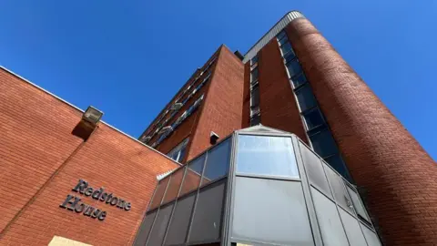 Henry Godfrey-Evans/BBC A brick-built block of flats with several storeys, with a glass entrance porch and "Redstone House" in silver lettering on a brick wall.