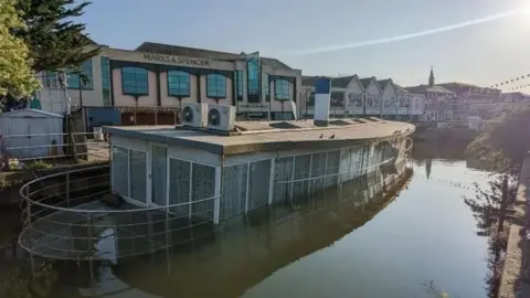 Ports of Truro A boat which is half submerged in water. Shops behind it.