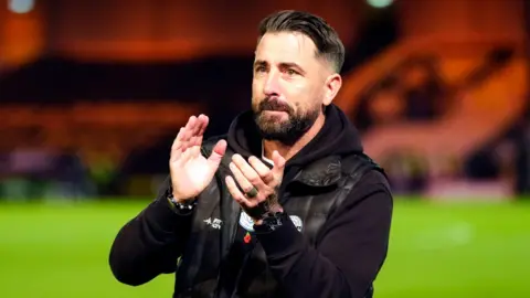 PA Media Kevin Horlock is wearing a black bodywarmer over a black hoodie. He has brown hair and a beard and is clapping on a football pitch.
