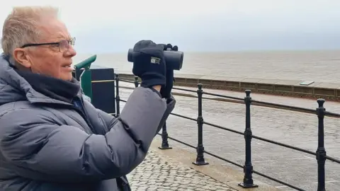 Stuart Baines A man with grey hair and black rimmed spectacles holds up a set of binoculars to his face. He is wearing a grey puffa jacket and standing on a cobbled pavement behind black metal railings looking out to sea. In the background is a road a low wall and a grey sea stretching to the horizon and a light blue sky.