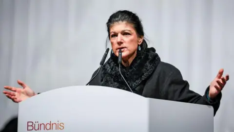 Leonhard Simon/Getty Images Standing on a stage, Wagenknecht launches her campaign behind a lectern wearing a large grey scarf