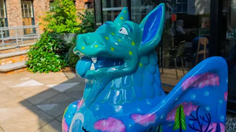 Make it York A sculpture of a mythical figure in blue and green. It stands on a paved area with buildings behind it