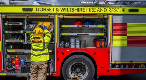DWFRS A fire truck with a firefighter putting an item into a shelf in the truck. It says "Dorset and Wiltshire Fire and Rescue" at the top of the vehicle.