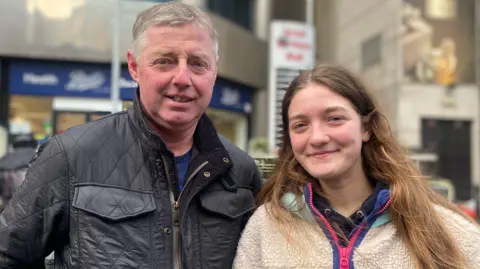 Alan is wearing a black jacket. He has grey hair. His daughter wears a wool fleece. She has long brown hair. There is a Boots store behind them.