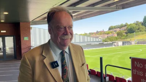 BBC/Peter Spencer Simon Myers has short grey hair and a grey moustache and is wearing a blue shirt with coloured tie and a brown jacket. Behind him is the football ground of Scarborough Athletic.