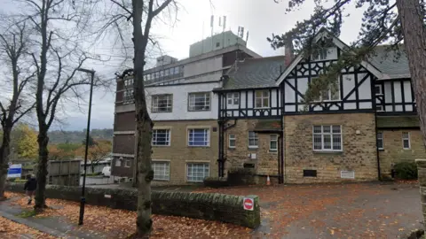 Google A large stone and brick building stands beside a tree-lined road on an overcast day. Part of the building features black-and-white timber framing in a Tudor-style design, while adjoining sections are constructed from light stone and brown brick. A taller modern building rises behind it, with antennas visible on the roof. Fallen autumn leaves cover the pavement and roadside, and several bare trees stand in the foreground. A low stone wall curves around the entrance area, where a red and white “no entry” sign is mounted.