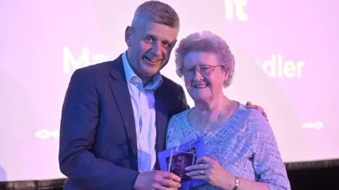 Park Christmas Savings Maureen Chandler stands next to Julian Coghlan at an award ceremony. Maureen has short grey hair and wears a long sleeved white sparkly top. She is holding her award in one hand. Julian is taller than Maureen and wears a dark blue suit jacket and white shirt. He is also holding Maureen's award in one hand.