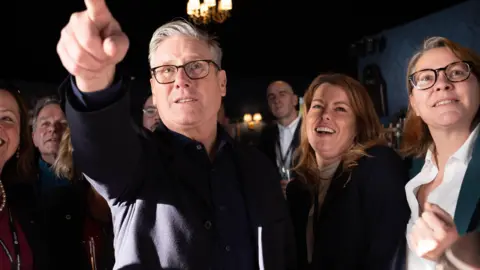 PA Media Keir Starmer pointing in front of him. He has grey, short hair and is wearing a dark suit and shirt with black glasses. Kim McGuinness is standing to the right and is laughing. She is wearing a dark jacket and beige top underneath. There are people around them looking in the same direction which smiles on their faces.