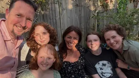 Mat Six people squeezing in for a selfie. Davina McCall is in the middle, she is surrounded by three young girls and their mum and dad. They are sitting in a garden.
