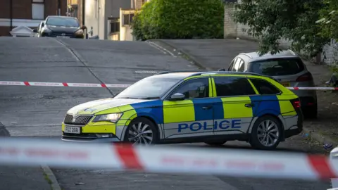 Aodhán Roberts A police car sits blocking a road. Either side are strips of police cordon. 
