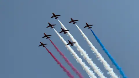 Eight red jet planes fly in a diamond formation across a blue, sunny sky. The aircraft are releasing coloured red, white and blue smoke which is trailing behind them.