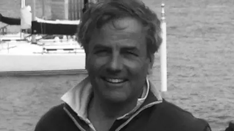 Ramsey Marina Ltd Headshot of Robin. He is smiling at the camera and stands on the coast - with the sea and a few boats behind him.