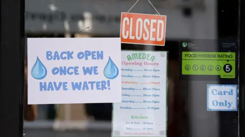 PA Media A sign on a glass door that says closed and another that reads "back open once we have water". Stickers detailing opening hours, a food hygiene rating and that the business is "card only" are also seen.