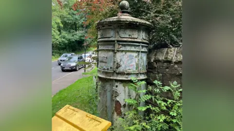 Northern Powergrid An electricity distribution pillar with peeling, faded green paint revealing a rusted base beneath. It stands against a sandstone weathered wall with overgrown shrubbery surrounding it.