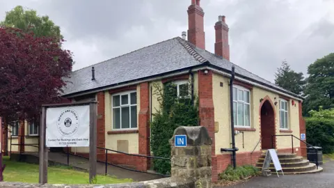 BBC/Carla Fowler The exterior of a small, single-storey community building situated in a quiet, green area. The building is constructed with cream-coloured textured walls and red brick detailing around the corners, windows, and doorways. The roof is covered with dark grey slate tiles and features multiple brick chimneys rising above it.
On the left side of the image, a large sign is mounted on a wooden frame. The sign clearly reads: “Newby and Scalby Community Hall” and underneath it says: “Contact for Bookings and Event Hire 01723 500699".